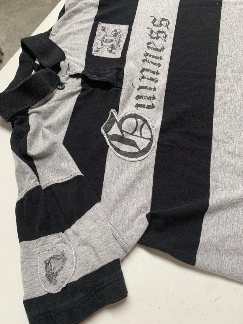 May include: A gray and black striped polo shirt with a black embroidered logo on the left chest and a black embroidered patch on the right sleeve. The logo says "Unlimited Sacrifice".