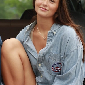 May include: A woman wearing a light blue denim shirt with a peace sign patch on the pocket. The patch is an American flag design. She is sitting in the back of a truck and has her legs crossed. She is wearing white trainers.