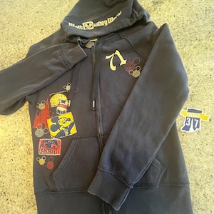 May include: A dark navy blue zip-up hoodie featuring the text Walt Disney World on the hood, the number 71, and various colorful graphic patches including Mickey Mouse and Animal Kingdom. The hoodie includes a front zipper and two pockets.