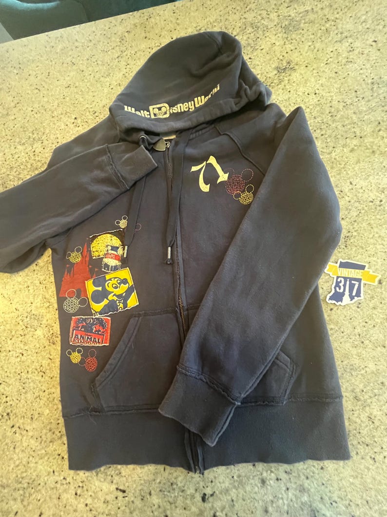 May include: A dark navy blue zip-up hoodie featuring the text Walt Disney World on the hood, the number 71, and various colorful graphic patches including Mickey Mouse and Animal Kingdom. The hoodie includes a front zipper and two pockets.