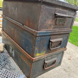 Op de afbeelding: Drie gestapelde metalen opbergdozen met handvatten. De bovenste doos is bruin, de middelste doos is blauw en de onderste doos is grijs. Elke doos heeft een nummer aan de voorkant: 14, 32 en 54.