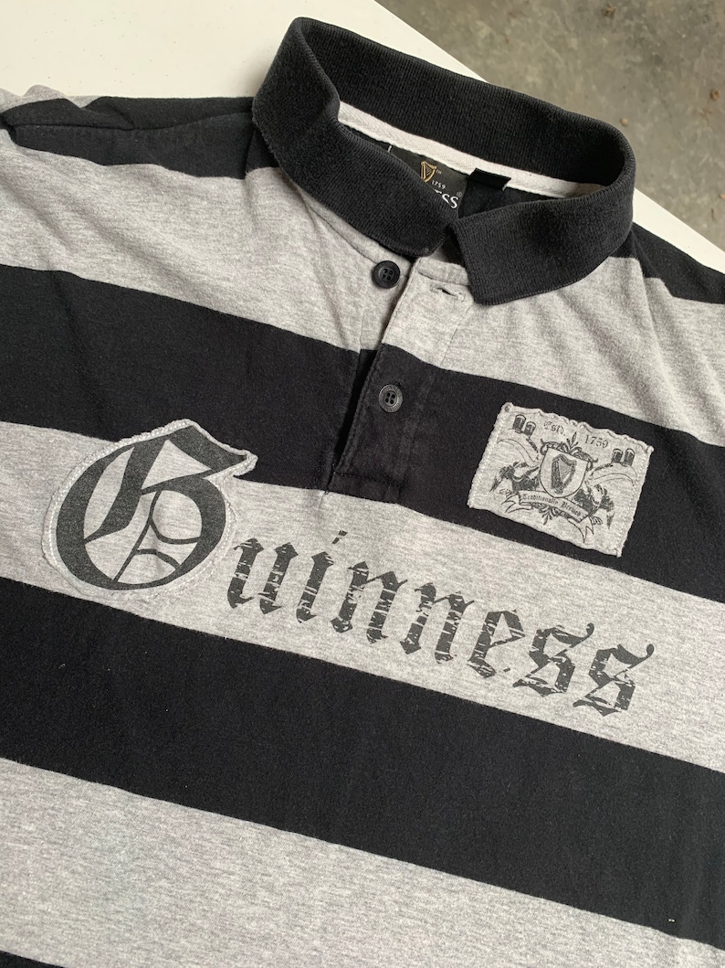 May include: A black and gray striped polo shirt with a large "Guinness" logo in a gothic font. The shirt also features a small patch with the Guinness logo and the text "Est. 1778" above it.