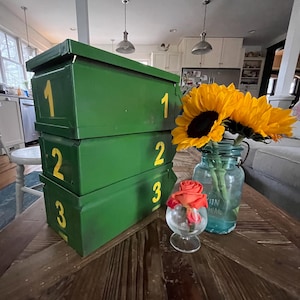 Op de afbeelding: Drie gestapelde groene metalen dozen met gele nummers 1, 2 en 3. Een boeket zonnebloemen in een blauwe glazen pot en een roos in een klein glas staan op een houten tafel. De dozen zijn rechthoekig.