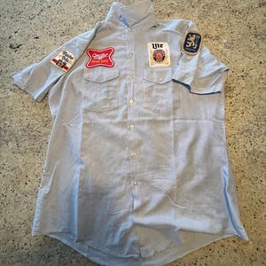 Może przedstawiać: Jasnoniebieska i biała koszula robocza w paski z krótkim rękawem i wieloma naszywkami. Naszywki to m.in. "Miller High Life", "Lite" i "Master Brew". Koszula ma dwie kieszenie na piersi i zapięcie na guziki.