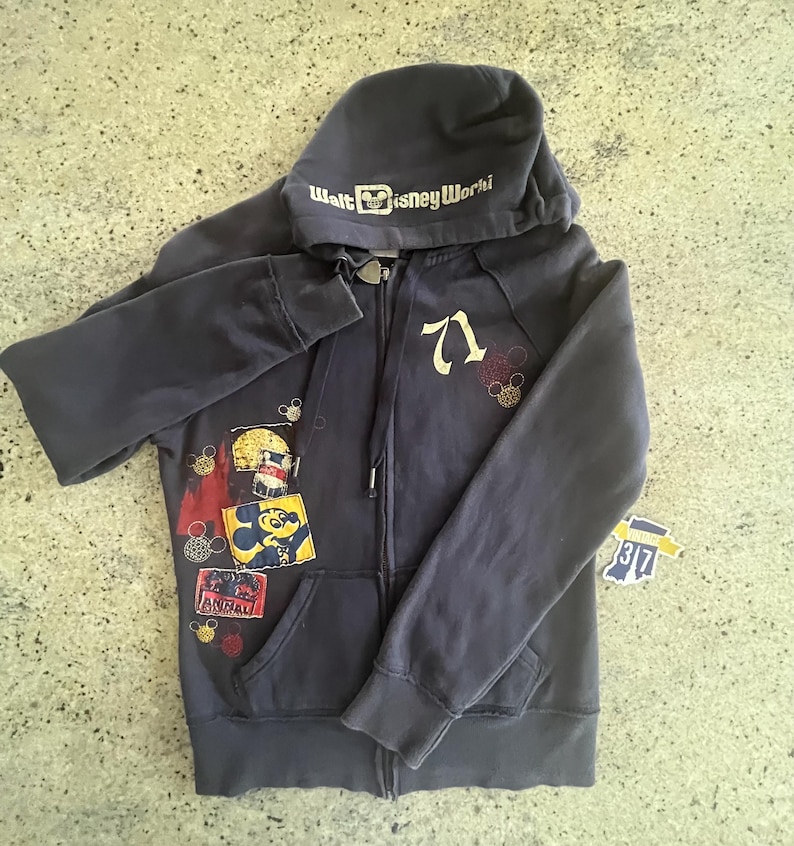 May include: A dark gray zip-up hoodie featuring the text Walt Disney World on the hood, the number 71 on the chest, and various colorful patches including a Mickey Mouse graphic. The garment includes long sleeves, a hood with drawstrings, and pockets.