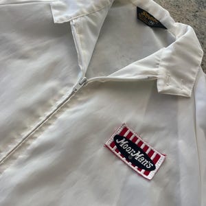 May include: White jacket with a zip fastening and a red and white striped patch with the text "Moor Mart's" on it. The jacket has a label that says "Sherwood" and "M" on it.