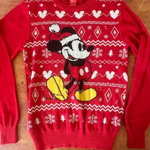Peut inclure: Pull-over rouge de Noël avec un motif de flocon de neige blanc et Mickey Mouse. Mickey Mouse porte un bonnet de Père Noël.