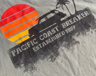 May include: A gray t-shirt with a graphic print of a sunset, a car, palm trees, and the text "Pacific Coast Breaker Established 1981".
