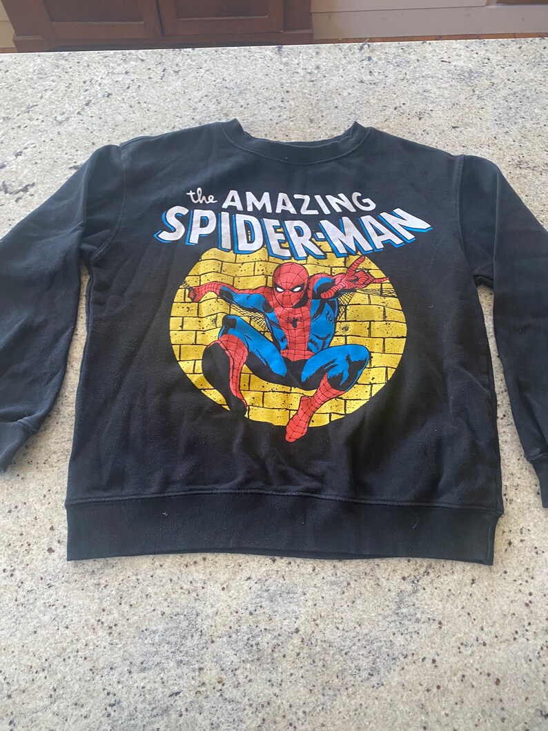 Puede incluir: Sudadera negra de manga larga con un gr&aacute;fico amarillo y rojo de Spider-Man. El texto "the AMAZING SPIDER-MAN" est&aacute; impreso en la parte delantera de la sudadera.