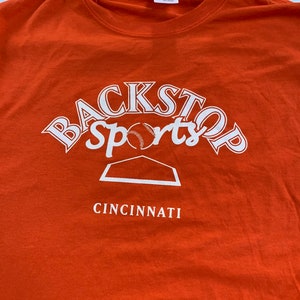May include: Orange t-shirt with a white graphic of a baseball diamond and the text "Backstop Sports Cincinnati"