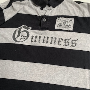 May include: A black and gray striped polo shirt with a large white "Guinness" logo and a small patch with the Guinness logo and the text "Est. 1778" on the right chest.