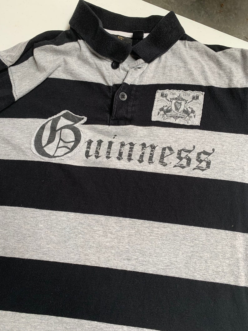 May include: A black and gray striped polo shirt with a large white "Guinness" logo and a small patch with the Guinness logo and the text "Est. 1778" on the right chest.