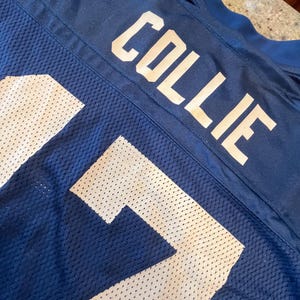 May include: A dark blue athletic jersey featuring the name COLLIE printed in bold white capital letters across the upper back. The garment is constructed from a breathable mesh fabric with a visible texture and a solid dark blue collar trim.