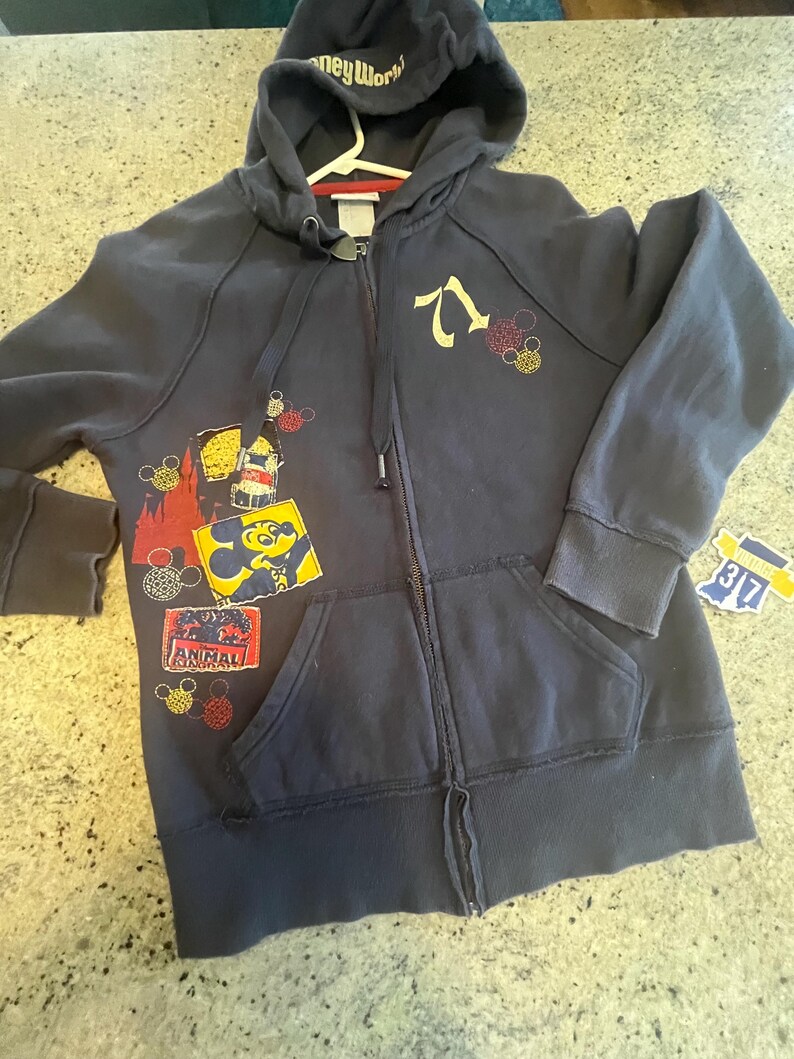 May include: A dark blue zip-up hoodie featuring the text Disney World on the hood, the number 71, and various graphic patches including a Mickey Mouse icon, a red castle silhouette, and an Animal Kingdom logo with small circular patterns.