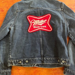 May include: A dark blue denim jacket with a red and white Miller High Life patch on the back.