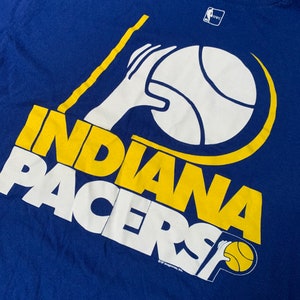 May include: Blue t-shirt featuring the Indiana Pacers logo in white and yellow. The logo includes a basketball, a hand, and the team name. The shirt has a crew neck and a vintage look.