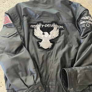 May include: Black leather jacket with a Harley-Davidson eagle patch on the back and a Harley-Davidson logo patch on the right sleeve. The jacket has a zipper closure and a ribbed waistband.