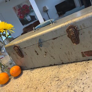 Puede incluir: Una caja de herramientas de metal vintage con un asa azul y pátina oxidada. La caja de herramientas tiene dos cierres y una pegatina en el lateral.