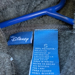 May include: Blue and white clothing tag with the text "Disney" and "S" for size. The tag also includes information about the fabric composition, care instructions, and origin of the garment. The tag is made of a blue fabric with white text.