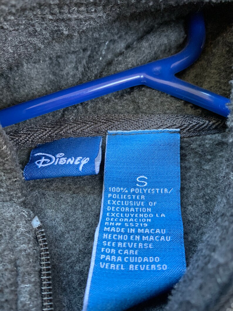 May include: Blue and white clothing tag with the text "Disney" and "S" for size. The tag also includes information about the fabric composition, care instructions, and origin of the garment. The tag is made of a blue fabric with white text.