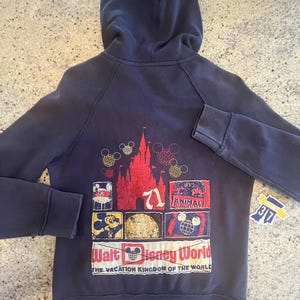 May include: A dark blue zip-up hoodie featuring a back graphic with a red castle, Mickey Mouse icons, and various park-themed patches. The text reads Walt Disney World, The Vacation Kingdom of the World, and Animal Kingdom, with a small tag showing 37.