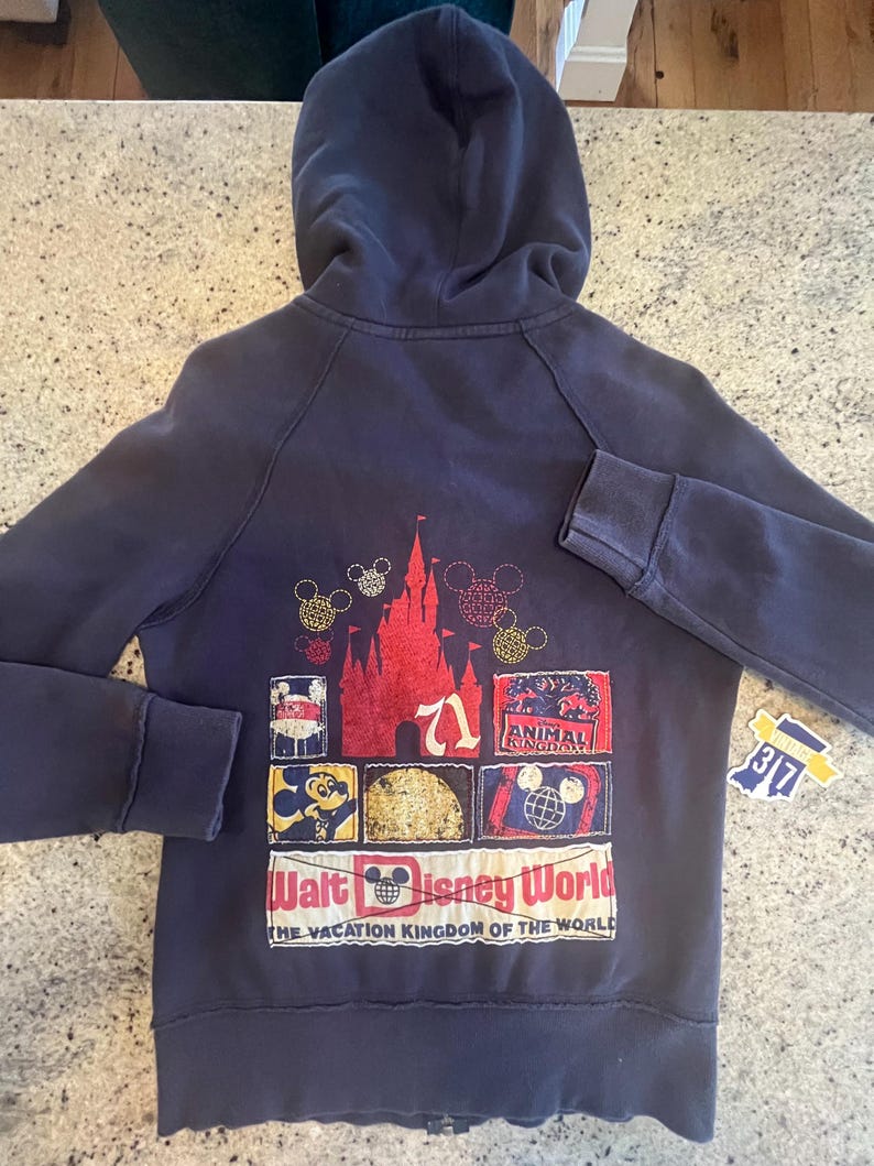 May include: A dark blue zip-up hoodie featuring a back graphic with a red castle, Mickey Mouse icons, and various park-themed patches. The text reads Walt Disney World, The Vacation Kingdom of the World, and Animal Kingdom, with a small tag showing 37.