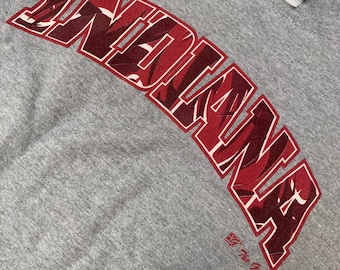 Könnte beinhalten: Graues T-Shirt mit einem roten "Indiana"-Motiv mit Tarnmuster. Das Motiv ist in einer großen, fetten Schrift gehalten und über die Vorderseite des Shirts gewölbt. Das Shirt hat auch ein kleines "The Game"-Logo unter dem Motiv.
