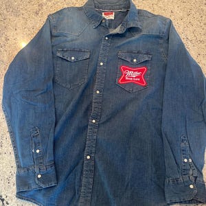 Puede incluir: Camisa vaquera de manga larga con cierre de botones, dos bolsillos en el pecho y puños abotonados. Un parche con el logotipo de Miller High Life en rojo y blanco está en el pecho izquierdo. La camisa es de un lavado azul medio.