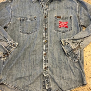 Può includere: Una camicia di jeans blu chiaro a maniche lunghe e con bottoni. La camicia ha una tasca a toppa sul petto sinistro con un logo "Miller High Life" rosso e bianco.