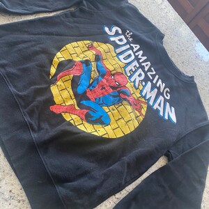 Puede incluir: Sudadera negra de manga larga con un estampado gr&aacute;fico de Spiderman en un c&iacute;rculo amarillo con el texto "the AMAZING SPIDER-MAN".