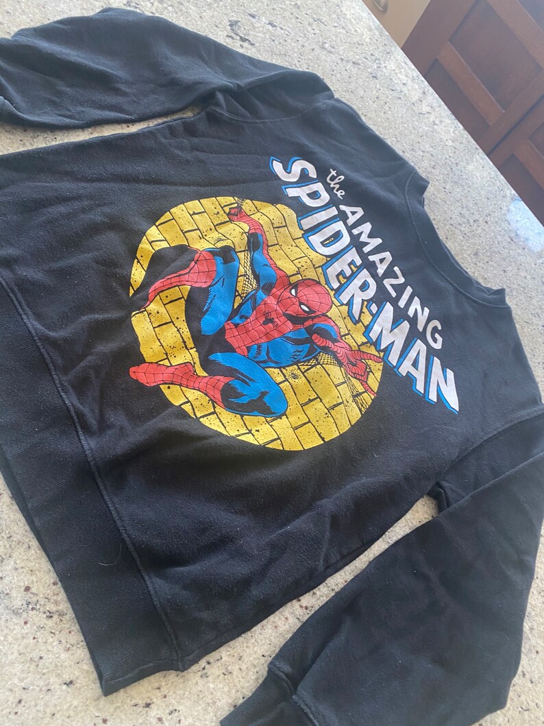 Puede incluir: Sudadera negra de manga larga con un estampado gr&aacute;fico de Spiderman en un c&iacute;rculo amarillo con el texto "the AMAZING SPIDER-MAN".