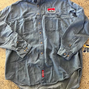 May include: A light blue denim long-sleeve shirt with a red "Marlboro" logo patch on the left chest. The shirt has two chest pockets and a button-down front.