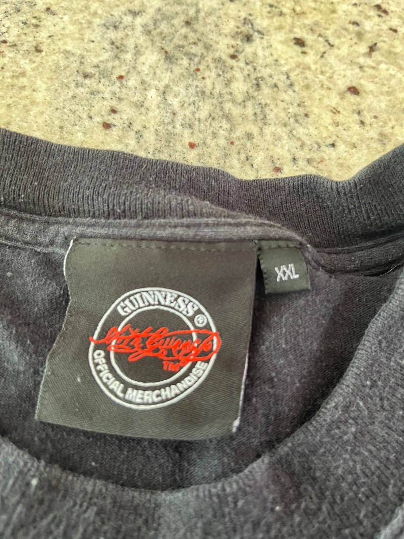 Puede incluir: Camiseta negra con una etiqueta negra que dice "GUINNESS" en letras blancas y "Official Merchandise" en letras blancas alrededor de un c&iacute;rculo rojo con una firma roja en su interior. La etiqueta tambi&eacute;n tiene un s&iacute;mbolo "TM" blanco. La etiqueta tiene una etiqueta de talla "XXL" blanca.