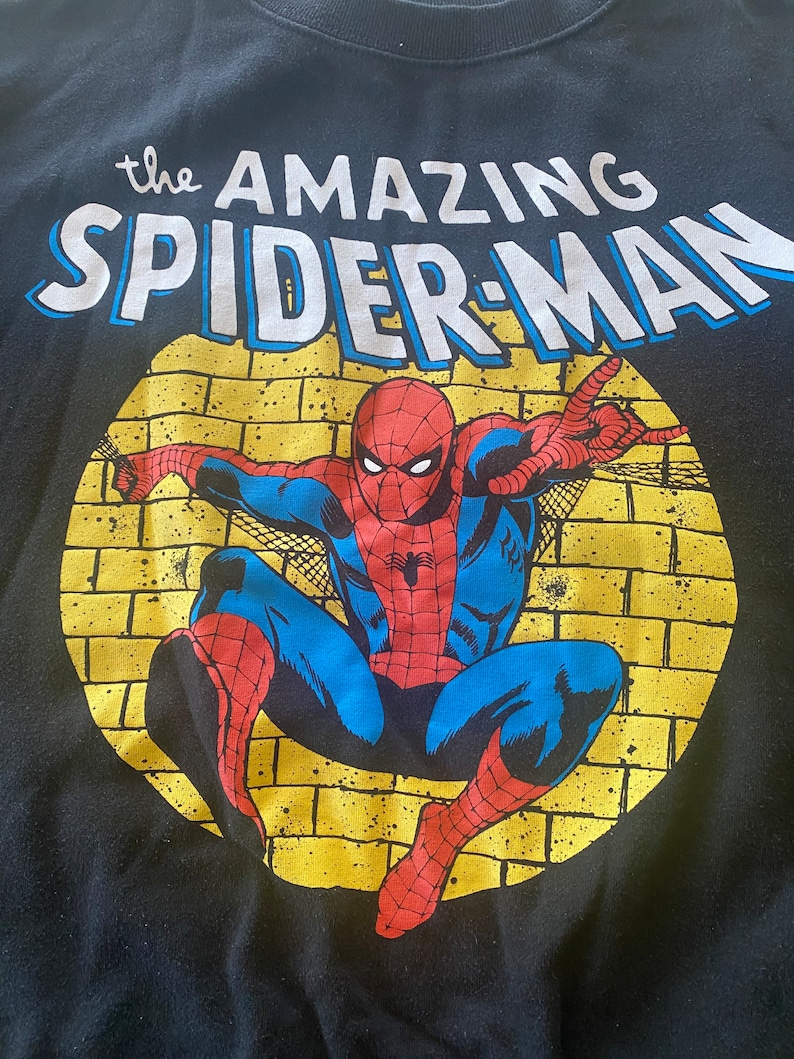 Puede incluir: Camiseta negra con un gr&aacute;fico circular amarillo que presenta un personaje de Spiderman rojo y azul en una pose de lanzamiento de telara&ntilde;a. El texto "the AMAZING SPIDER-MAN" est&aacute; impreso sobre el gr&aacute;fico.