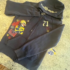 May include: A dark navy blue zip-up hoodie featuring the text Walt Disney World on the hood. The front displays the number 71 in gold, along with colorful patches including a Mickey Mouse graphic and an Animal Kingdom logo with small icons.