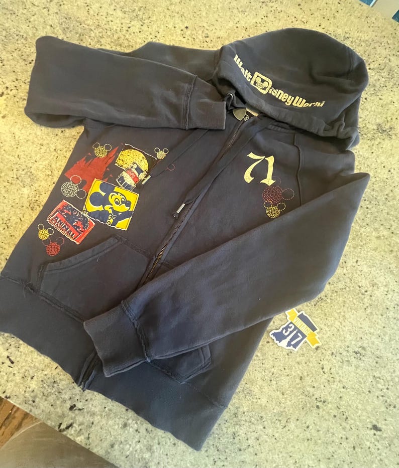 May include: A dark navy blue zip-up hoodie featuring the text Walt Disney World on the hood. The front displays the number 71 in gold, along with colorful patches including a Mickey Mouse graphic and an Animal Kingdom logo with small icons.