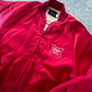 May include: A vibrant red satin bomber jacket with a zip-up front. The jacket has a white and red Miller High Life logo patch on the chest. The collar and cuffs are a darker shade of red.