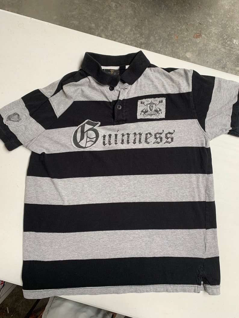 May include: A black and gray striped polo shirt with the word "Guinness" printed on the front. The shirt has a small patch with a crest on the right side of the chest.