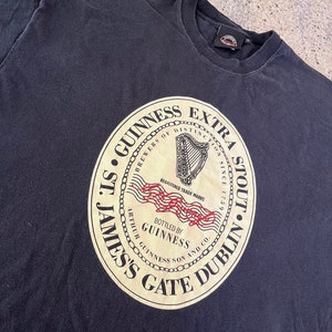 Puede incluir: Camiseta negra con un gr&aacute;fico circular de un sello dorado con el texto "St. James's Gate Dublin", "Guinness", "Brewers of Distinction", "Extra Stout" y "Bottled by Guinness Arthur Guinness Son and Co."