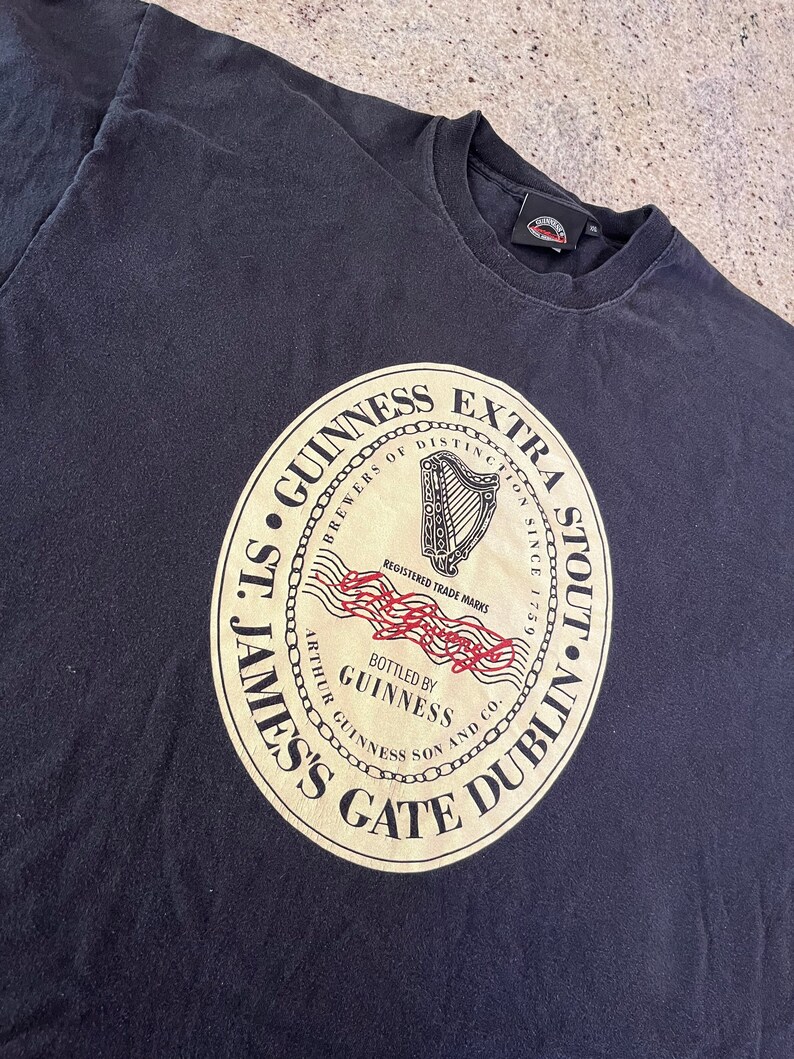 Puede incluir: Camiseta negra con un gr&aacute;fico circular de un sello dorado con el texto "St. James's Gate Dublin", "Guinness", "Brewers of Distinction", "Extra Stout" y "Bottled by Guinness Arthur Guinness Son and Co."
