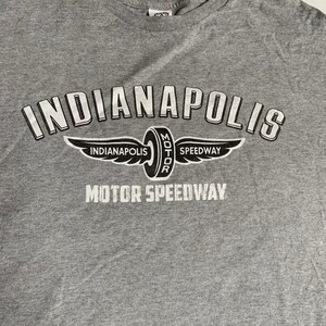 Może przedstawiać: Szara koszulka z białym nadrukiem logo Indianapolis Motor Speedway. Logo przedstawia oponę z napisem "Indianapolis Motor Speedway" i skrzydlatym wzorem.
