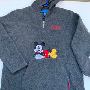 May include: A gray fleece pullover with a half-zip closure and a kangaroo pocket. The pullover features a Mickey Mouse graphic on the pocket and the word "MICKEY" embroidered on the chest.