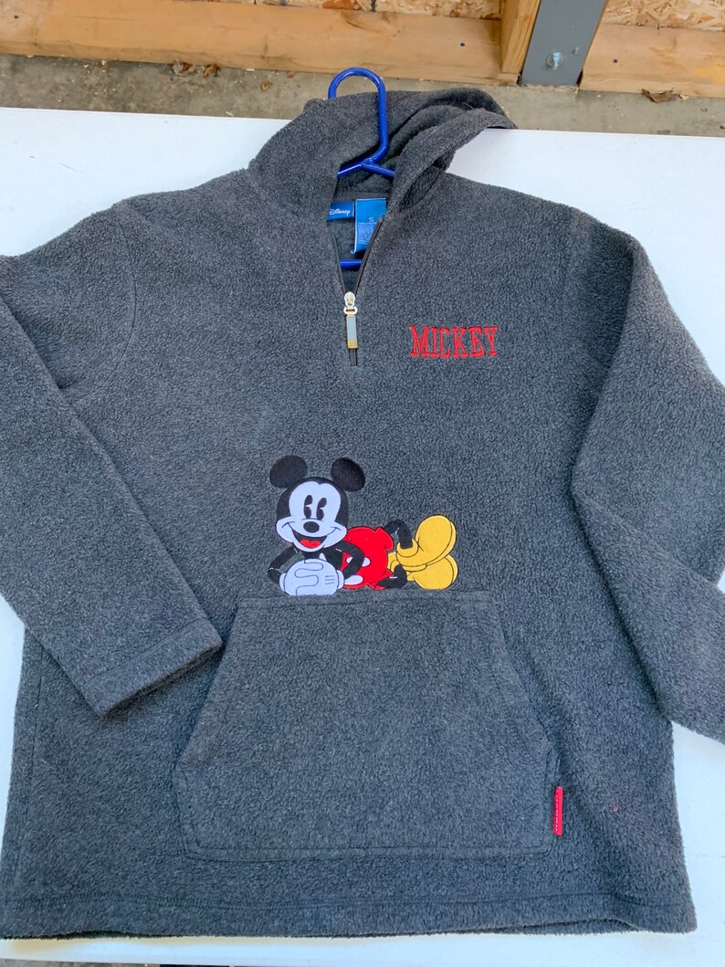May include: A gray fleece pullover with a half-zip closure and a kangaroo pocket. The pullover features a Mickey Mouse graphic on the pocket and the word "MICKEY" embroidered on the chest.