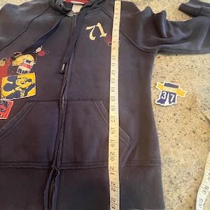 May include: A dark blue zip-up hoodie featuring a graphic of Mickey Mouse and the number 71. A white measuring tape is placed vertically along the front, showing a length of approximately 24 inches from the shoulder to the bottom hem.