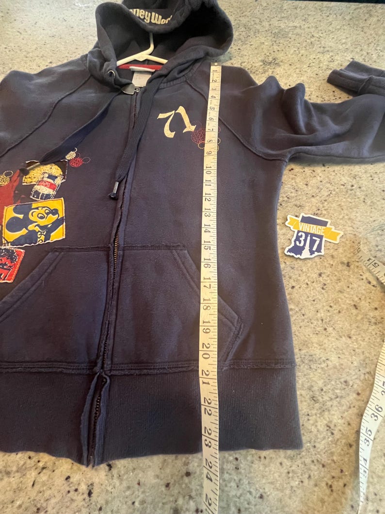 May include: A dark blue zip-up hoodie featuring a graphic of Mickey Mouse and the number 71. A white measuring tape is placed vertically along the front, showing a length of approximately 24 inches from the shoulder to the bottom hem.
