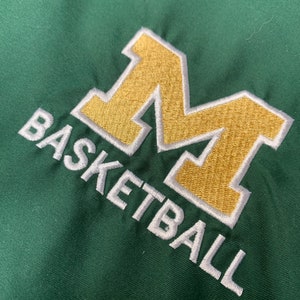 Puede incluir: Un parche bordado en verde y dorado con la letra "M" y la palabra "BASKETBALL" debajo.