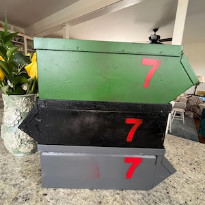 Op de afbeelding: Drie gestapelde metalen dozen, groen, zwart en grijs geverfd. Elke doos heeft een rood nummer 7 op de voorkant. De dozen zijn in een piramidevorm gerangschikt. De dozen lijken handgemaakt.