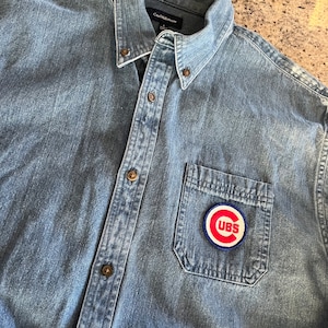 May include: A blue denim shirt with a red, white, and blue Chicago Cubs patch on the left chest pocket. The shirt has a button-down collar and a button closure. The brand name "Croft & Barrow" is visible on the inside collar.