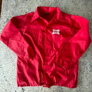 May include: A vibrant red jacket featuring a button-up front, a collar, and two front pockets. A white and red Miller logo is on the left chest. The jacket is made of a shiny, lightweight material.