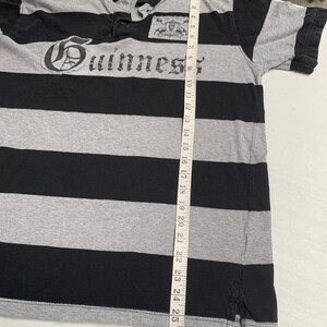 May include: A black and gray striped polo shirt with a Guinness logo on the front. The shirt has a button-down collar and short sleeves.